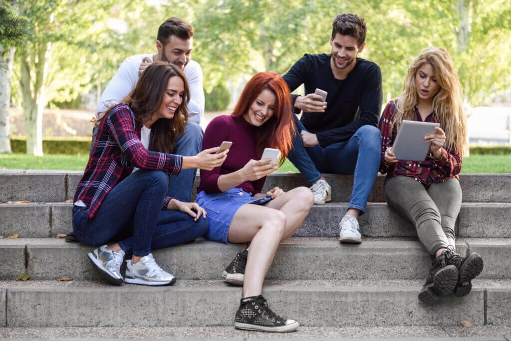 CoCarting wishlist app image: A group of five young adults sitting on outdoor steps, laughing and looking at their smartphones and a tablet, engaged in a shared digital experience.